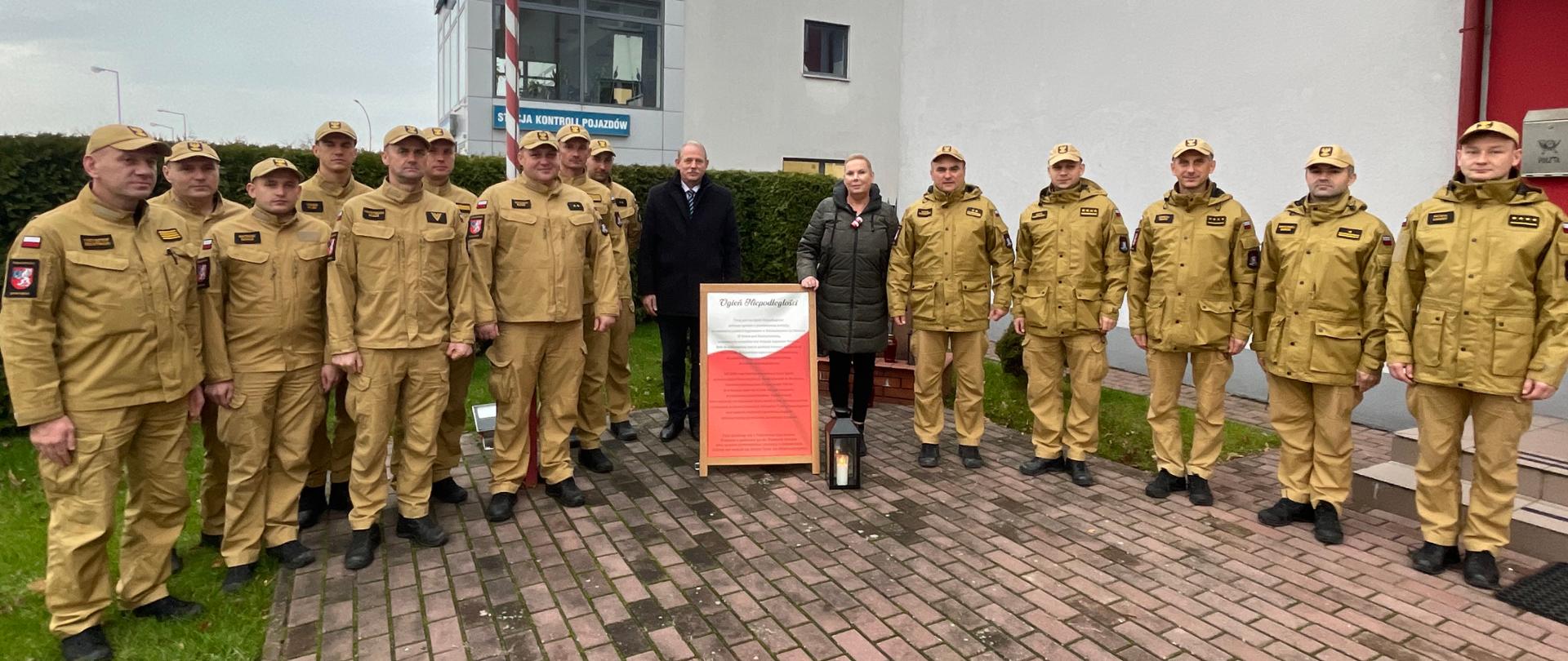 Ujęcie, koncentrujące się na grupie osób przed fragmentem białego budynku straży pożarnej. Na pierwszym planie, centralnie, znajduje się kobieta w ciemnej kurtce trzymająca w dłoni tablicę "Ogień Niepodległości". Po jej lewej i prawej stronie stoi kilkunastu mężczyzn w beżowo-żółtych mundurach polowych i czapkach. Za kobietą i częścią mężczyzn, bliżej budynku, stoi dwóch mężczyzn w ciemnych garniturach/płaszczach. Grupa pozuje do zdjęcia w zwartej formacji. Tablica, czarna latarnia z płomieniem oraz mundur oficerów stanowią główne elementy kompozycji, podkreślając uroczysty charakter spotkania związany z przekazywaniem symbolicznego ognia.