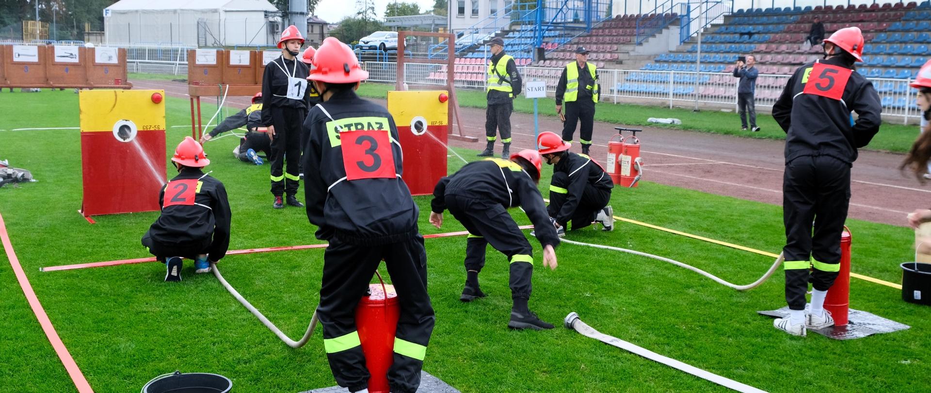 Na zdjęciu strażacy młodzieżowej drużyny pożarniczej podczas zawodów sportowo-pożarniczych. W tle sędziowie konkurencji oraz trybuny boiska.
