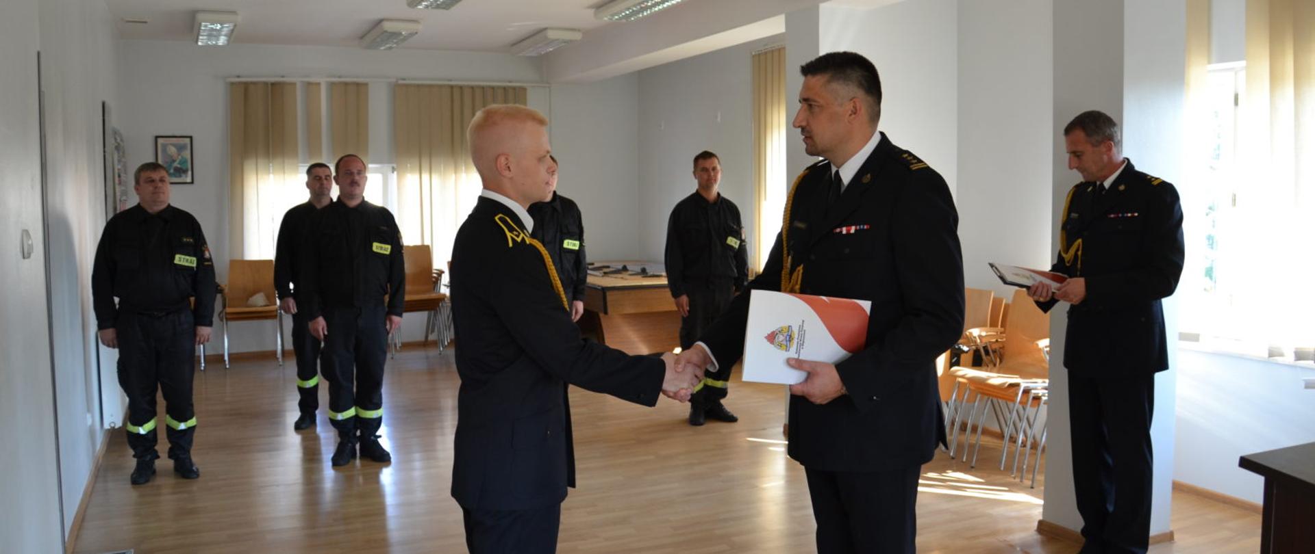 starszy brygadier Witold Wiercioch Komendant Powiatowy Państwowej Straży Pożarnej w Ropczycach stoi w postawie zasadniczej prawą dłonią ściska dłoń awansowanego na pierwszy stopień w korpusie aspiranckim młodszego aspiranta Tomasza Rumak, w lewej dłoni komendant trzyma akt nadania