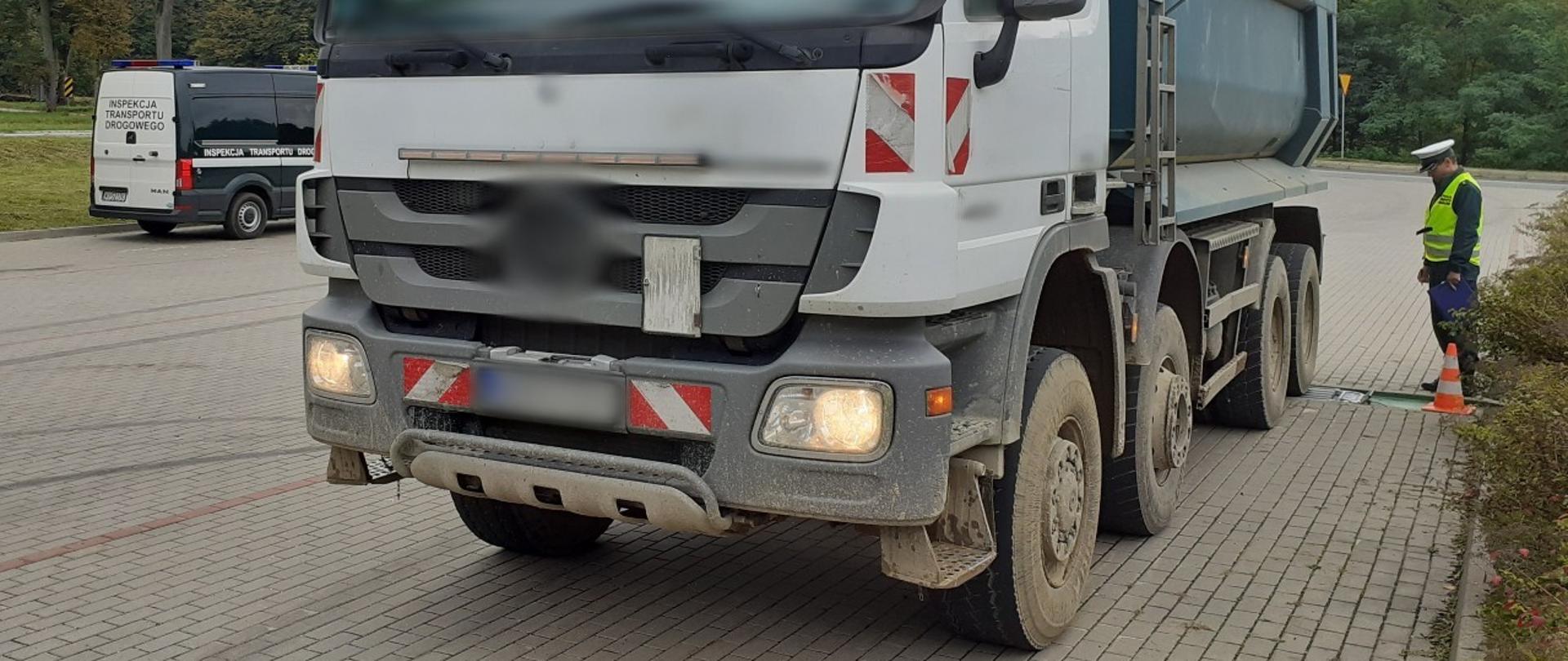 Na pierwszym planie: przód i lewy bok przeładowanej wywrotki z piachem. W tle: ciężarówka stoi kołami ostatniej osi na przenośnych wagach. Umundurowany inspektor warmińsko-mazurskiej ITD odczytuje wynik pomiaru z przenośnych wag.