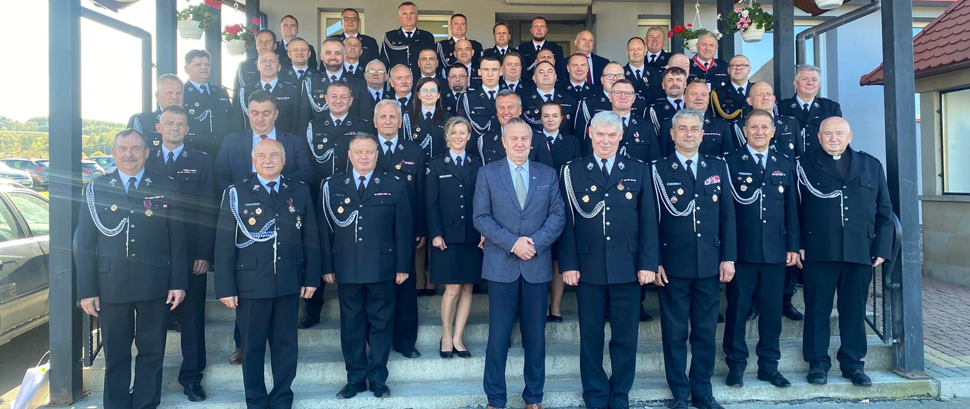 Uczestnicy VI Zjazdu Oddziału Powiatowego ZOSP RP w Rzeszowie pozują do wspólnego zdjęcia na schodach przed budynkiem Domu Strażaka w Harcie. Na fotografii znajduje się kilkudziesięciu przedstawicieli Państwowej Straży Pożarnej, Ochotniczych Straży Pożarnych, ZOSP RP oraz zaproszonych gości. Większość osób ubrana jest w ciemnogranatowe mundury galowe z odznaczeniami i sznurami galowymi. W pierwszym rzędzie stoją między innymi przedstawiciele władz strażackich oraz samorządowych. Nad uczestnikami znajduje się drewniane zadaszenie ozdobione wiszącymi donicami z kwiatami.