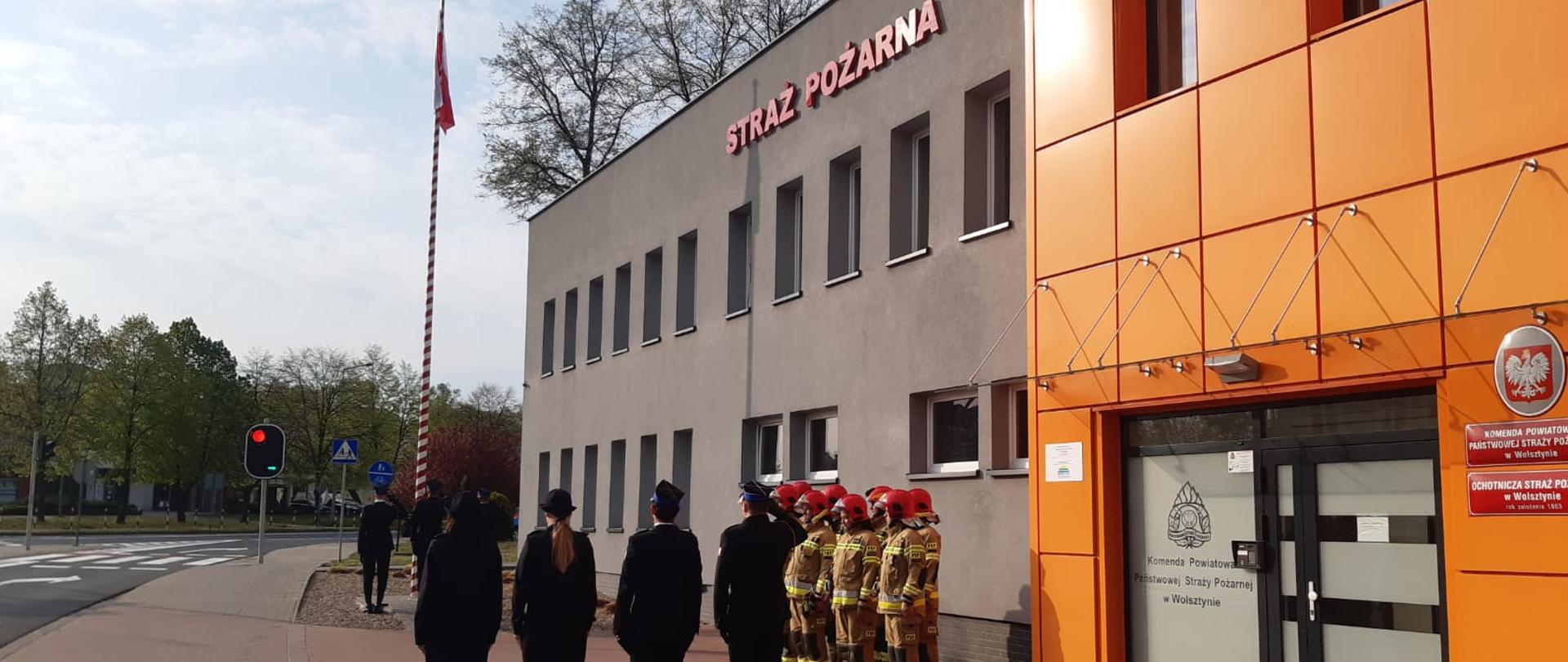 Strażacy w mundurach stoją przed budynkiem komendy przygotowani do uroczystości wciągnięcia flagi RP na maszt