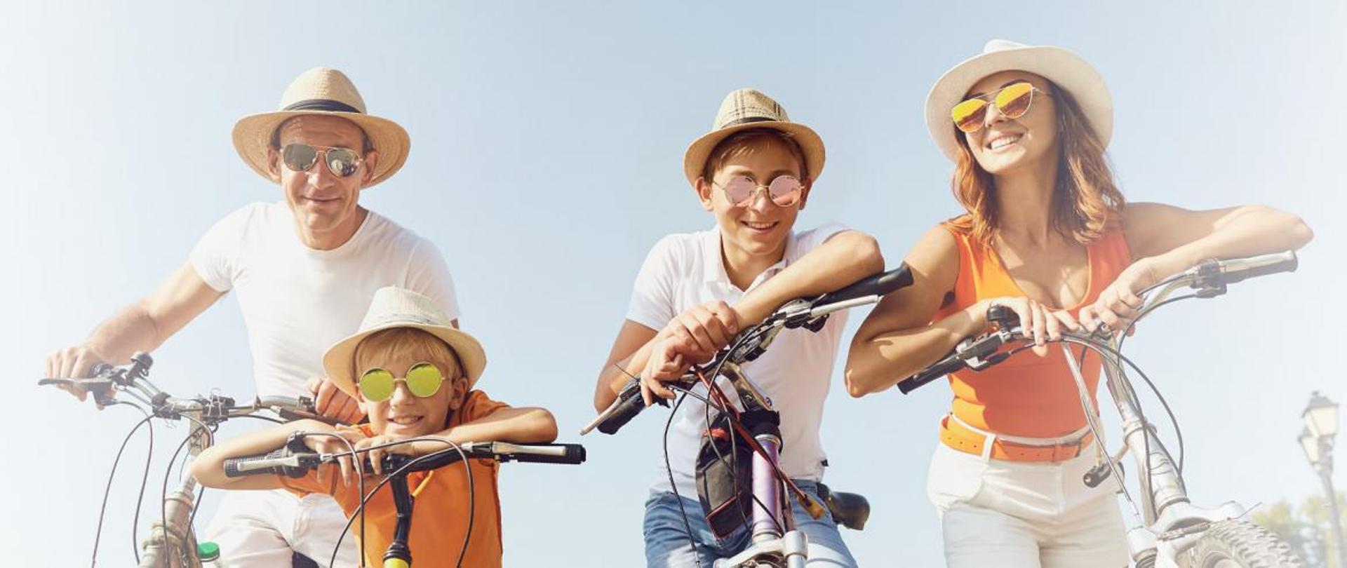 Na fotografii widoczna jest czteroosobowa rodzina. Wszyscy pozują do zdjęcia siedząc na rowerach. Każdy jest uśmiechnięty, ubrany w jasny kapelusz i przeciwsłoneczne okulary.