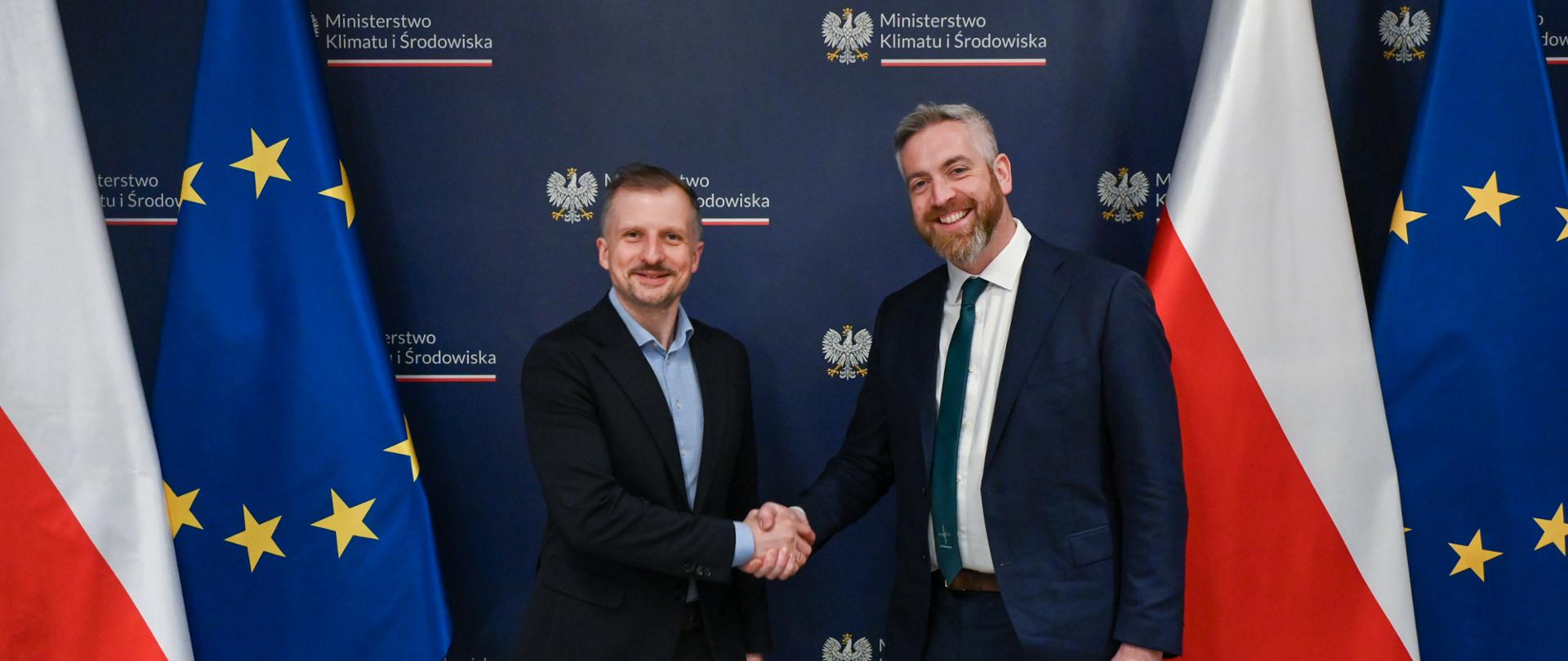 Wiceminister klimatu i środowiska Mikołaj Dorożała z wiceministrem Christopherem O’Sullivanem z Irlandzkiego Ministerstwa Budownictwa, Samorządów Lokalnych i Dziedzictwa. Wiceministrowie podają ręce i pozują na tle flag PL i UE.