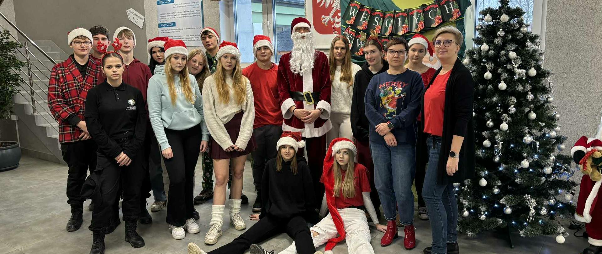 Młodzież w czapkach Mikołaja pozuje do wspólnego zdjęcia w świątecznej scenerii, stojąc obok przebranej za Mikołaja osoby i ozdobionej choinki. Cała grupa, w tym dwie nauczycielki, znajduje się na korytarzu szkolnym udekorowanym na Boże Narodzenie.