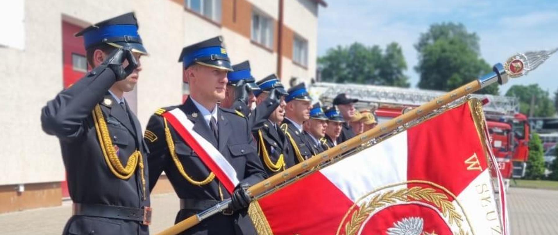 Funkcjonariusze straży pożarnej w umundurowaniu wyjściowym w szyku. Jeden salutuje, drugi trzyma pochylony sztandar.