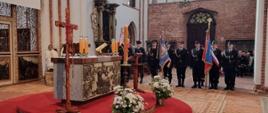 Wnętrze kościoła o wysokim sklepieniu i gotyckiej architekturze. Na podwyższeniu z czerwonym dywanem znajduje się ołtarz z krzyżem, świecami oraz dekoracjami kwiatowymi. Obok stoi ambona. Po prawej stronie w nawie głównej ustawiony jest poczet sztandarowy – kilku umundurowanych strażaków trzyma sztandary i stoi w szeregu. W tle widoczne są ceglane ściany, wysokie okna wpuszczające światło oraz elementy wyposażenia świątyni.