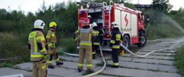 Widok z tyłu. Na pierwszym planie kilku strażaków OSP w ubraniach specjalnych stoi za samochodem pożarniczym w trakcie ćwiczeń.