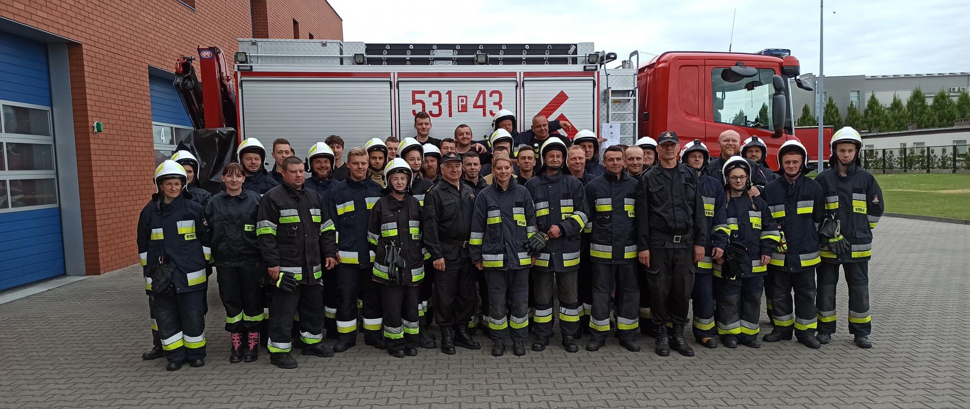 Zdjęcie grupowe uczestników w mundurach strażackich na tle samochodu pożarniczego.