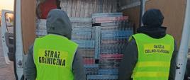 Dwóch funkcjonariuszy w kamizelkach z napisami służb stoi tyłem do aparatu i ogląda kartony umieszczone w otwartym busie.