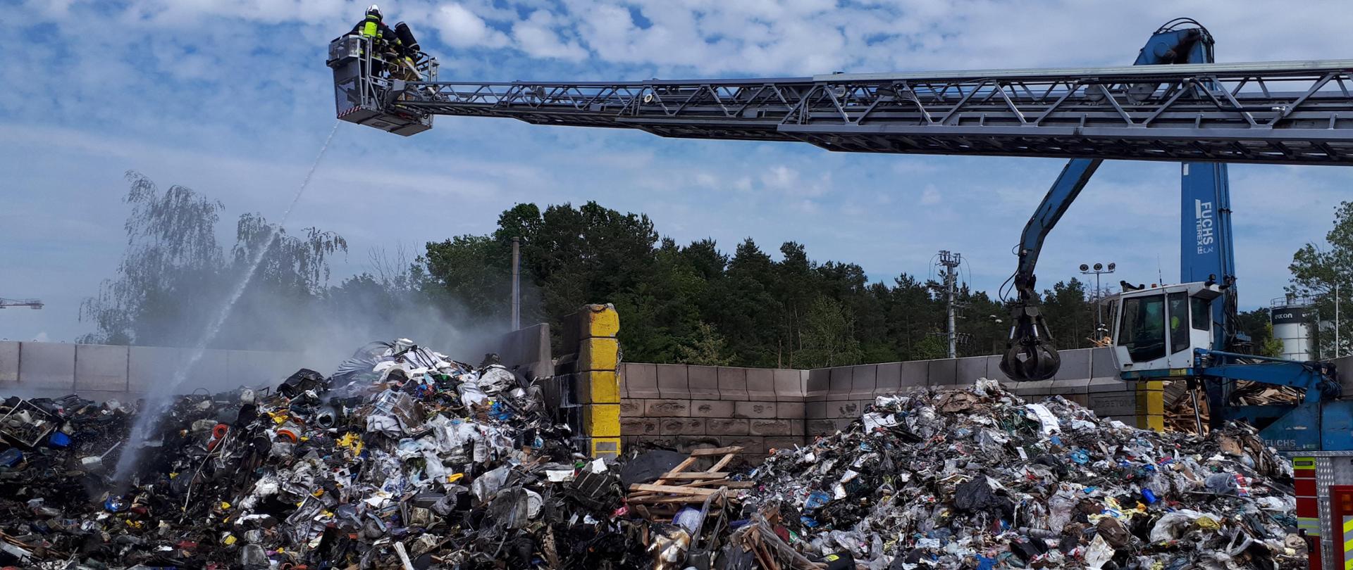 Pryzmy częściowo spalonych odpadów znajdują się na utwardzonym podłożu. Kilka metrów nad nimi, na stalowym wysięgniku, znajdują się dwaj funkcjonariusze Straży Pożarnej, którzy przelewają te odpady strumieniem wody. W tle widać betonowy płot ograniczający miejsce składowania odpadów. 