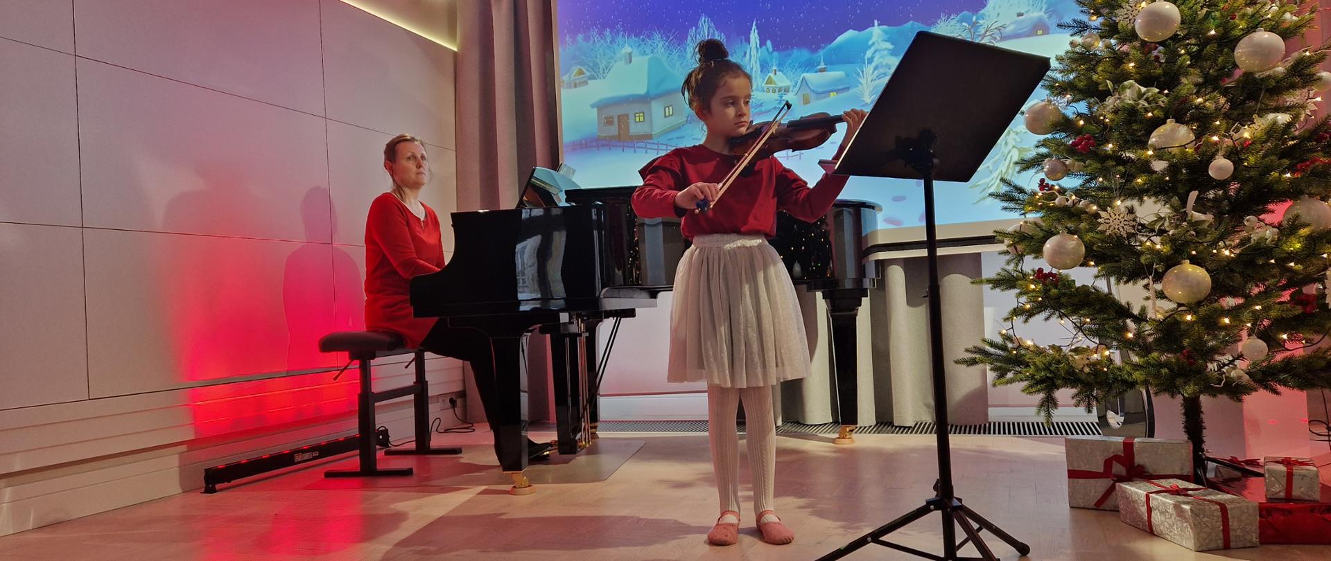 Zdjęcie przedstawia uczennicę stojącą na scenie i grającą na skrzypcach. Dziewczynka ubrana jest w czerwoną bluzkę i jasnoszarą spódniczkę. Przed uczennicą jest ustawiony czarny pulpit do nut. Za uczennicą z lewej strony stoi fortepian, przy którym siedzi nauczyciel ubrany w czerwoną bluzkę. Z prawej strony stoi choinka z prezentami. W tle na ekranie wyświetlona jest grafika, przedstawiająca zimowy krajobraz z ośnieżonymi domami i choinkami. Po bokach na ścianach i częściowo na suficie odbija czerwone światło. 