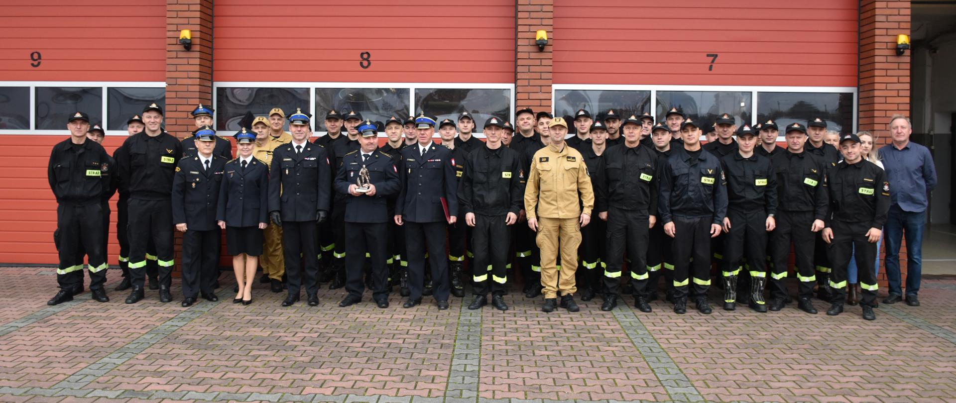 Zdjęcie przedstawia funkcjonariusza odchodzącego na zaopatrzenie emerytalne wraz z kolegami i koleżankami z pracy na wspólnym zdjęciu przed garażami JRG. 