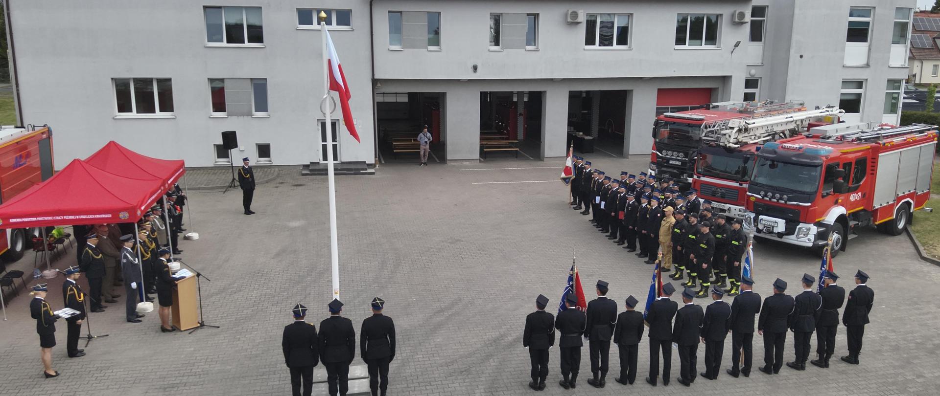 Fotografia przedstawia ujęcie placu apelowego wykonane z wysokości kilku metrów nad ziemią. Widoczny cały plac, po prawej stronie kadru stoją pododdziały pożarnicze, za ich plecami stoją pojazdy pożarnicze. W centralnej części kadru, tyłem do fotografującego stoją w szyku poczty towarzyszące i poczet flagowy przy maszcie z wciągniętą flagą państwową. Po lewej stronie kadru widoczna trybuna honorowa. Całość ujęcia na tle budynku Komendy Powiatowej PSP w Strzelcach Krajeńskich.