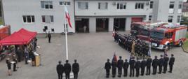 Fotografia przedstawia ujęcie placu apelowego wykonane z wysokości kilku metrów nad ziemią. Widoczny cały plac, po prawej stronie kadru stoją pododdziały pożarnicze, za ich plecami stoją pojazdy pożarnicze. W centralnej części kadru, tyłem do fotografującego stoją w szyku poczty towarzyszące i poczet flagowy przy maszcie z wciągniętą flagą państwową. Po lewej stronie kadru widoczna trybuna honorowa. Całość ujęcia na tle budynku Komendy Powiatowej PSP w Strzelcach Krajeńskich.