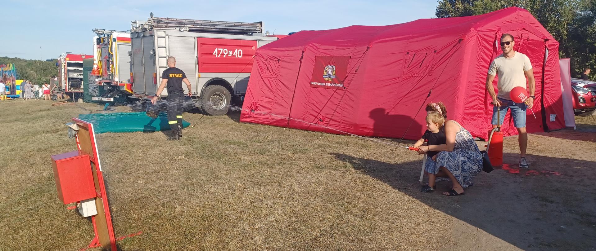 Zdjęcie przedstawia stoisko promocyjne komendy powiatowej państwowej straży pożarnej w Lipnie. Na zdjęciu rodzina, która pompuje wodę i celuje strumień do nalewaka