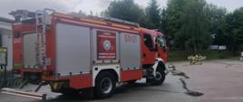 Na parkingu przy sklepie stoi samochód pożarniczy, w oddali stoją strażacy