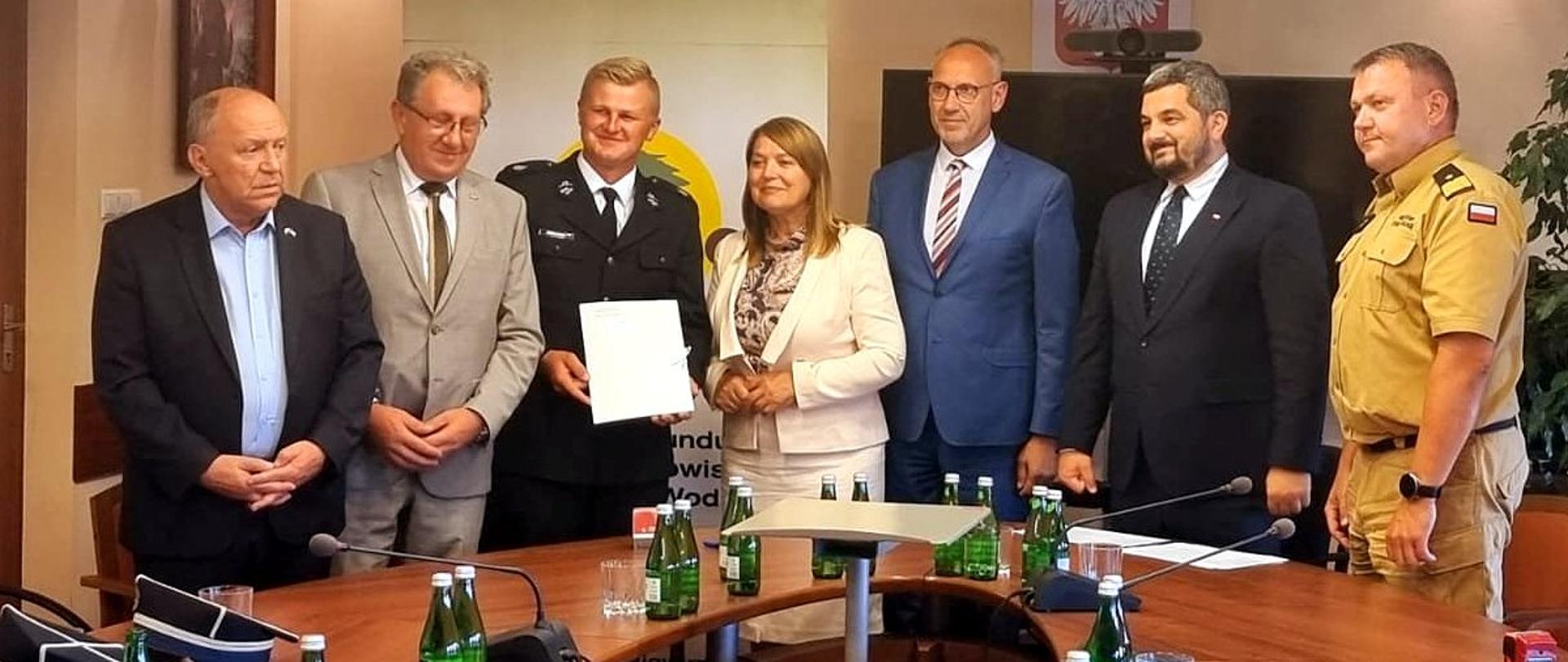 Zdjęcie zrobione w sali konferencyjnej. Przy stole konferencyjnym stoi 7 osób uczestniczących w uroczystości, wśród nich oficer Państwowej Straży Pożarnej w stopniu generała.