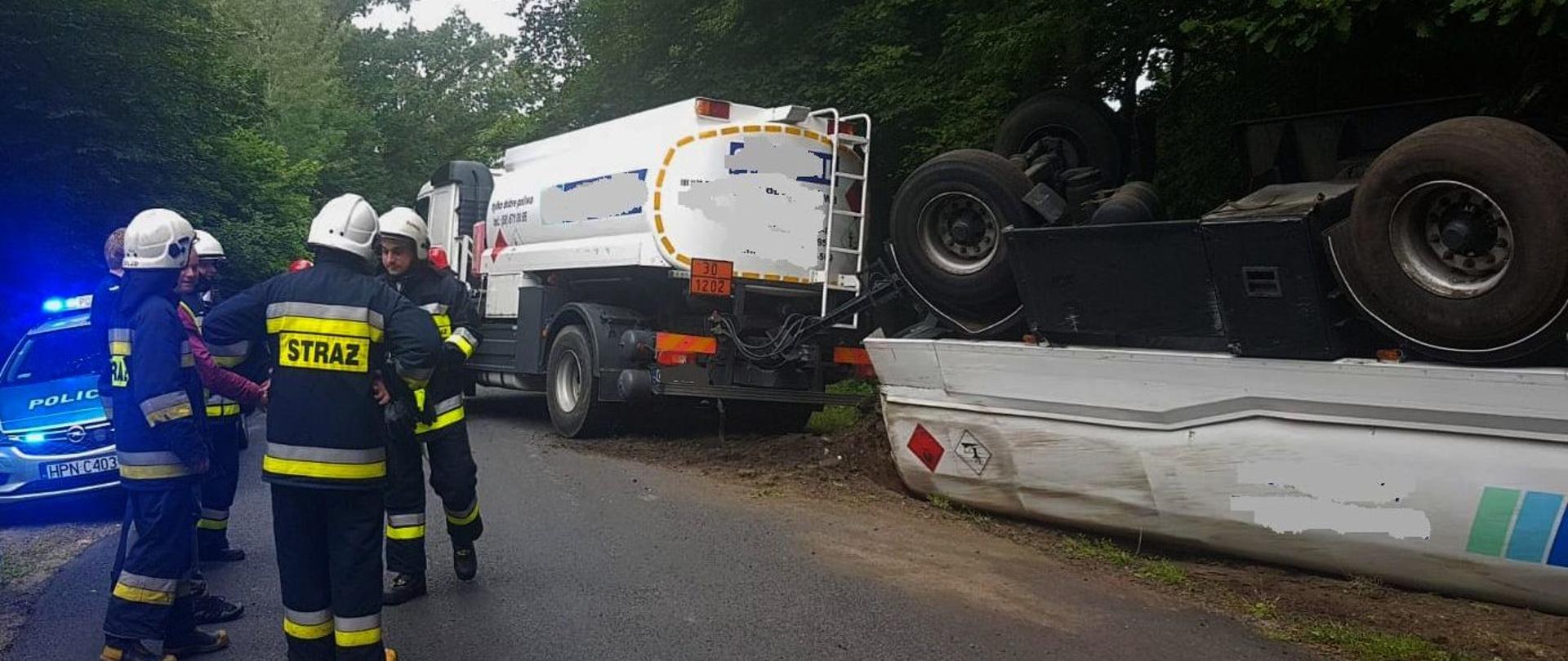 Zdjęcie przedstawia wypadek. Na pierwszym planie stoi grupa strażaków uczestniczących w działaniach, w tle widać białą ciężarówkę, do której podłączona jest naczepa. Naczepa znajduje się na poboczu, do góry kołami.
