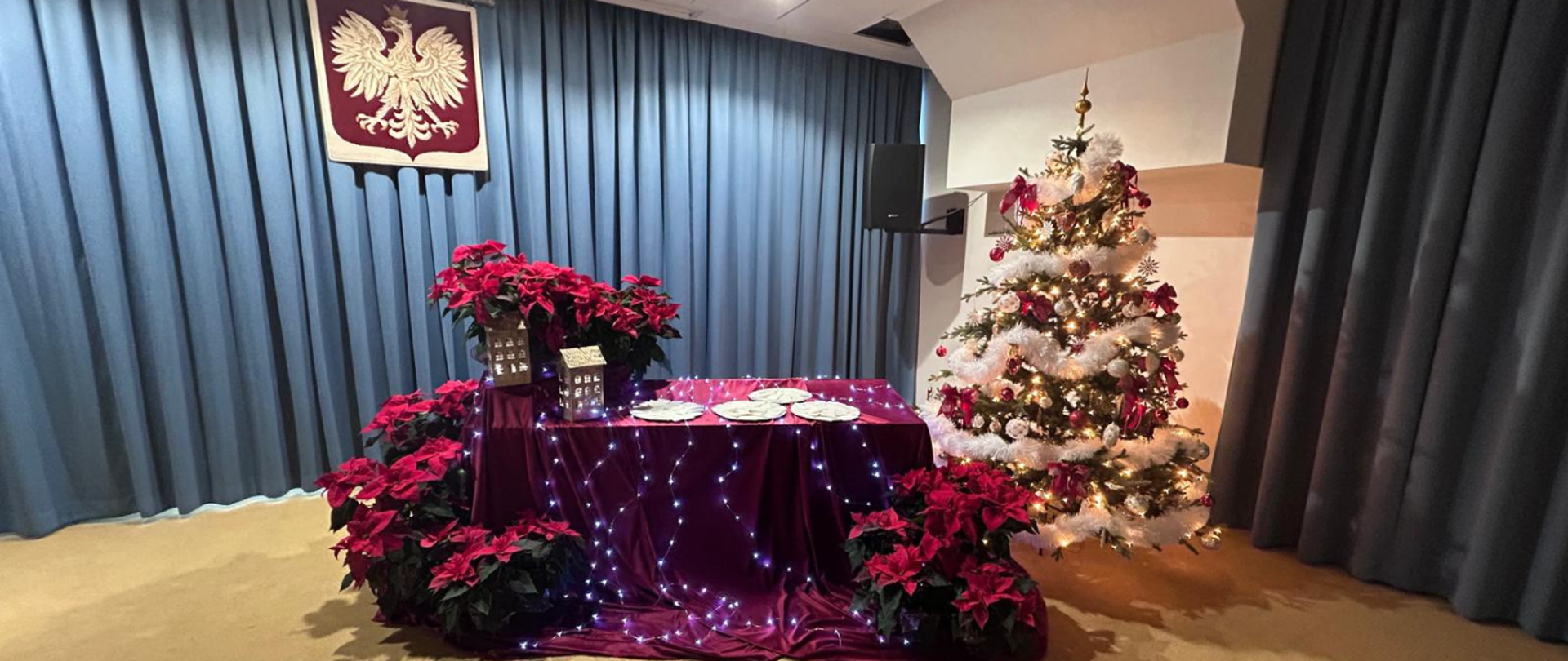 Na zdjęciu widoczny jest stół przystrojony bordowym obrusem i światełkami, na którym stoją lampiony, gwiazdy betlejemskie oraz talerze z opłatkami. Za stołem widoczna jest choinka z białymi i czerwonymi dekoracjami. W pomieszczeniu widoczne kremowe ściany, niebieska kotara oraz beżowa wykładzina. 