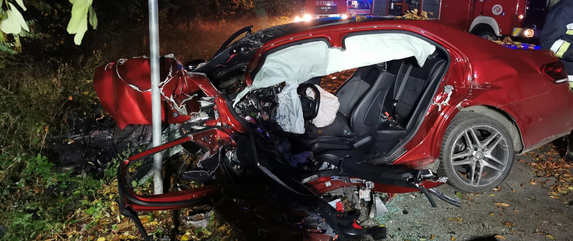 Rozbity samochód osobowy marki Mercedes stojący w poprzek jezdni, przodem przy znaku drogowym. W tle zastępy straży pożarnej i ZRM. 