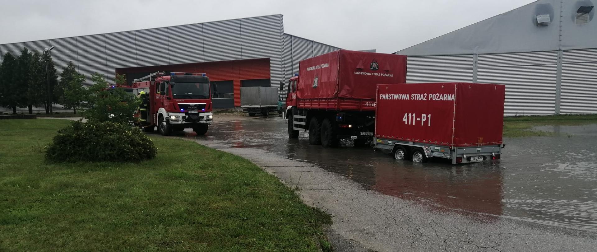 Zdjęcie przedstawia dwa samochody ratowniczo-gaśnicze. Za jednym z nich znajduję się przyczepka z pompą dużej wydajności. Za samochodami znajdują się dwie hale, obok nich zalega woda.