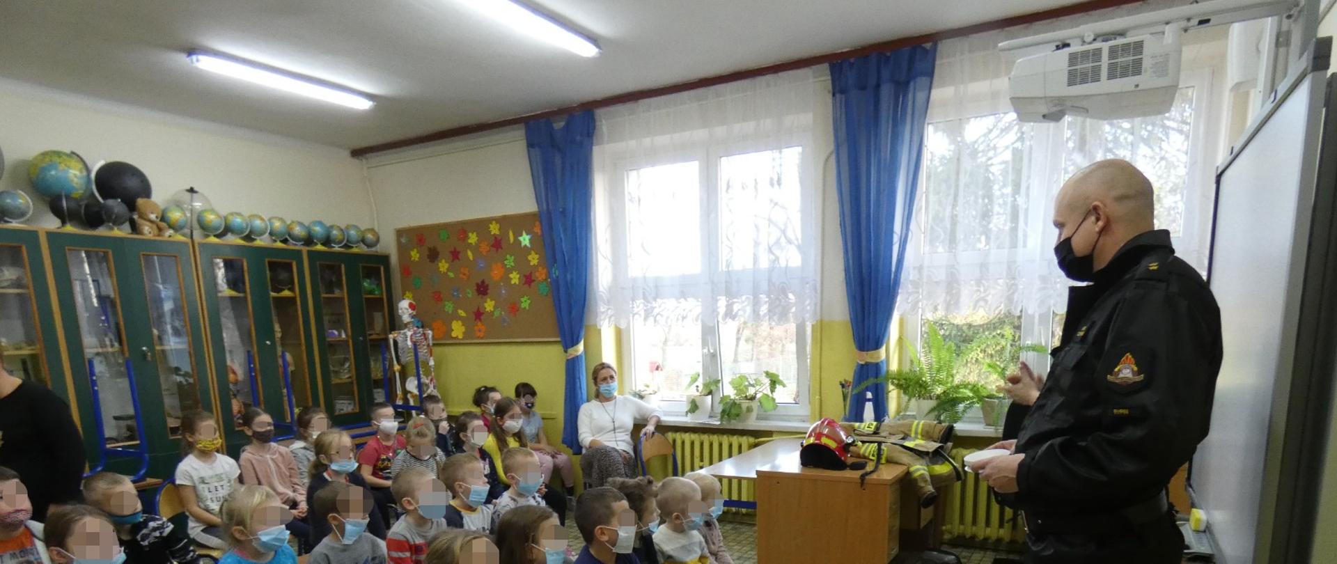 Zdjęcie wykonane jest w sali lekcyjnej i przedstawia strażaka, który przemawia do dzieci. Dzieci siedzą na ławeczkach, wśród nich jest Pani Nauczyciel. Za dziećmi w sali znajdują się szafki. W tle zdjęcia widać okna oraz biurko, na którym leży ubranie specjalne strażaka oraz hełm. Wszyscy mają założone na twarzy maseczki chirurgiczne. 