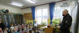 Zdjęcie wykonane jest w sali lekcyjnej i przedstawia strażaka, który przemawia do dzieci. Dzieci siedzą na ławeczkach, wśród nich jest Pani Nauczyciel. Za dziećmi w sali znajdują się szafki. W tle zdjęcia widać okna oraz biurko, na którym leży ubranie specjalne strażaka oraz hełm. Wszyscy mają założone na twarzy maseczki chirurgiczne. 