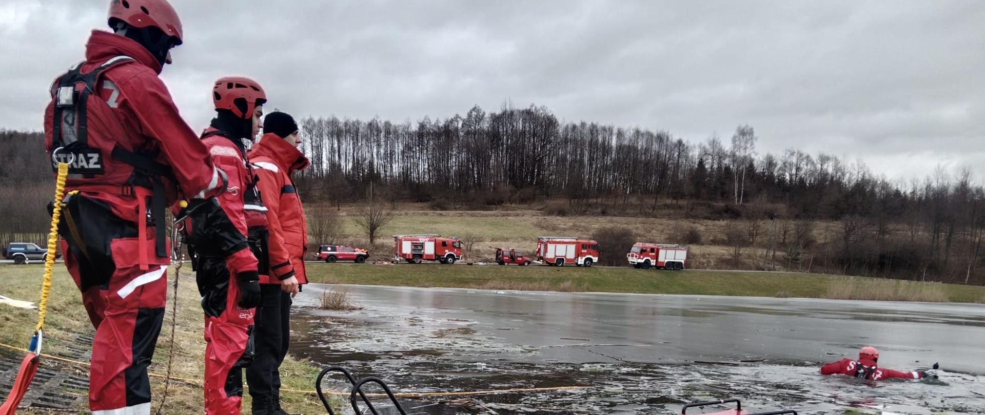 Kolorowa fotografia w pochmurny dzień wykonana na zalodzonym akwenie. Przedstawia strażaka znajdującego się w wodzie ubraniu w specjalnym zabezpieczony liną, trzech strażaków znajdujących się na brzegu również w ubraniach specjalnych oraz sanie lodowe. W tle samochody pożarnicze.
