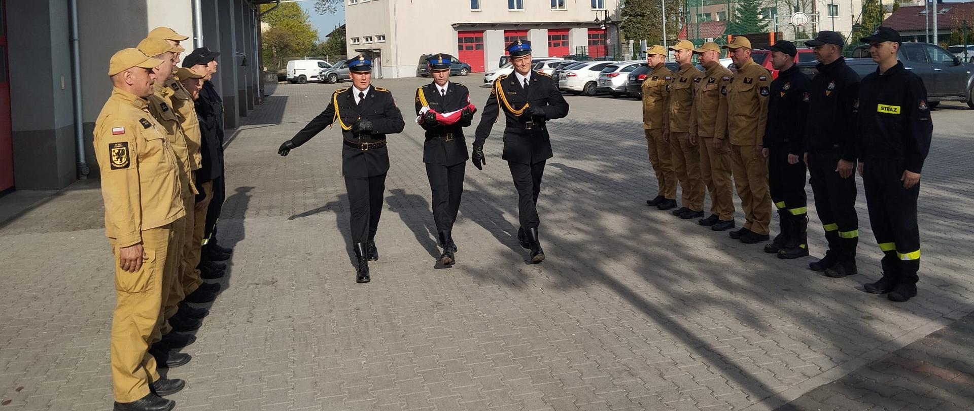 Poczet flagowy złożony z trzech strażaków maszeruje. Strażak na środku trzyma złożoną flagę państwową. Przed nimi strażacy poszczególnych zmian służbowych stojący naprzeciwko siebie w trakcie uroczystego wciągnięcia flagi państwowej na maszt.