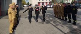 Poczet flagowy złożony z trzech strażaków maszeruje. Strażak na środku trzyma złożoną flagę państwową. Przed nimi strażacy poszczególnych zmian służbowych stojący naprzeciwko siebie w trakcie uroczystego wciągnięcia flagi państwowej na maszt.