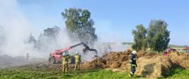 Widok na miejsce akcji. Na środku rota strażaków OSP podaje pierwszy prąd gaśniczy. Po lewej stronie strażak KP PSP podaje drugi prąd wody. Po prawej stronie przemieszczający się pieszo strażak OSP. Z tyłu maszyna rolnicza wspomagająca działania strażaków. W tle pola uprawne. 