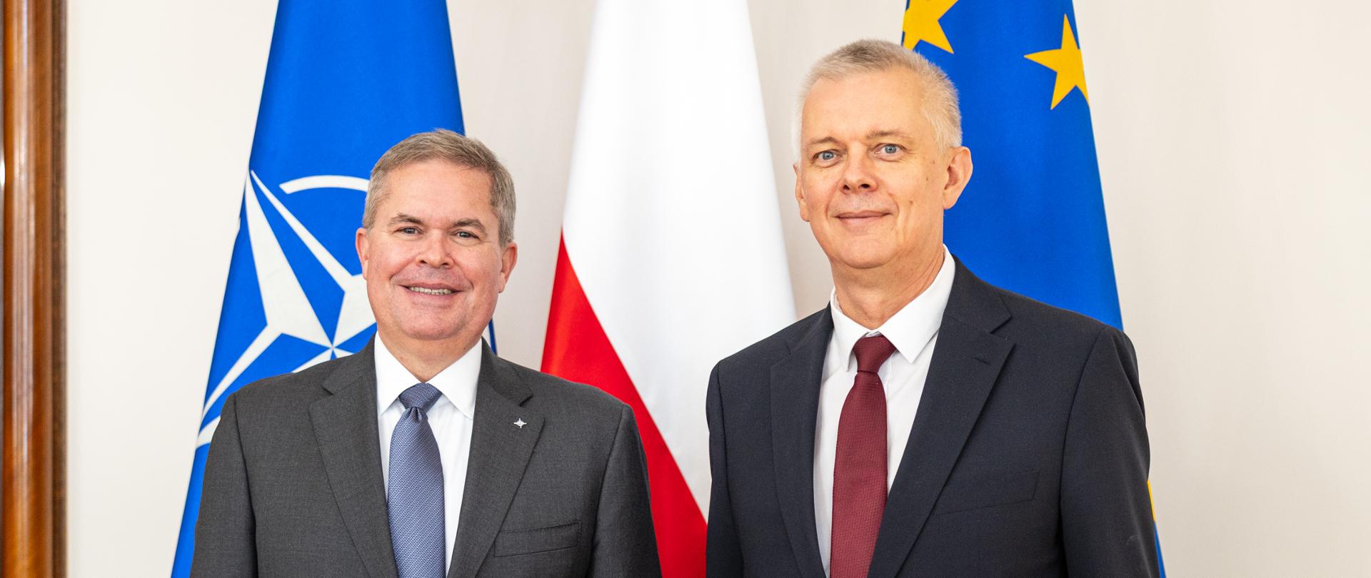 Na zdjęciu minister Tomasz Siemoniak (po prawej) oraz Scott W. Bray, zastępca sekretarza generalnego NATO do spraw wspólnego wywiadu i bezpieczeństwa (po lewej). Osoby stoją na tle trzech flag – w środku flaga Polski (biało-czerwona), po prawej flaga Unii Europejskiej (niebieska z żółtymi gwiazdkami) oraz po lewej flaga NATO (niebieska z białym logo). Po lewej stronie zdjęcia widać fragment drewnianych drzwi.
