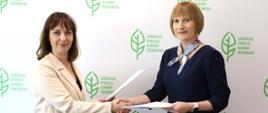 Na tle białej ściany z logotypem Generalnej Dyrekcji Ochrony Środowiska (zielony liść) stoją dwie kobiety. Kobiety ściskają sobie dłonie, a w drugiej ręce trzymają teczki. 