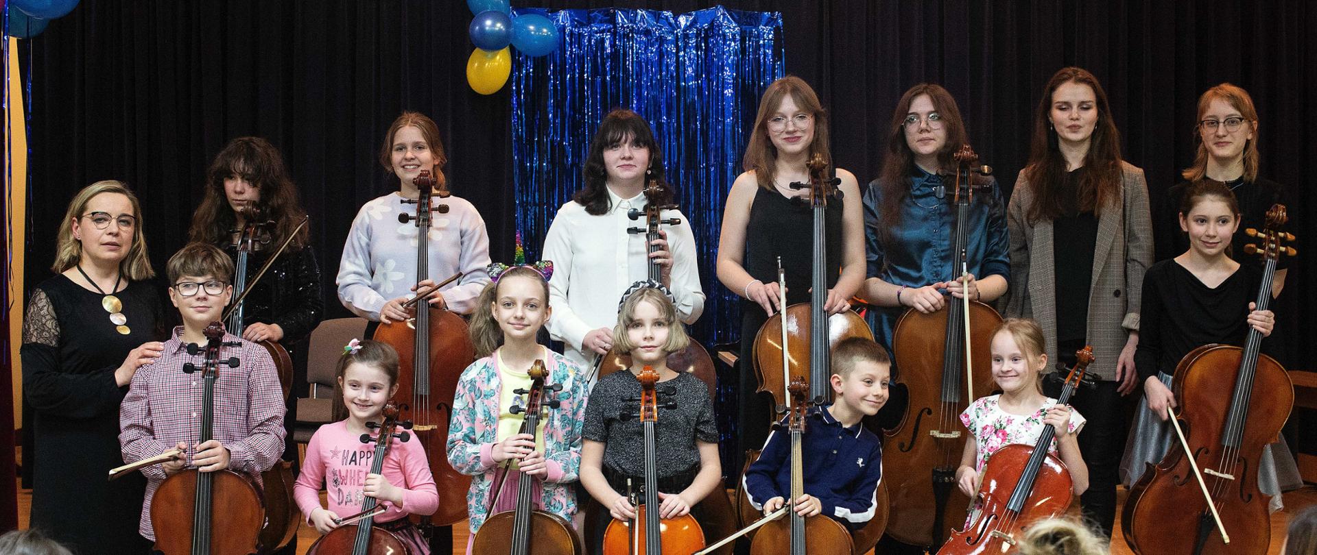 Zdjęcie przedstawia grupę uczniów stojących na scenie z wiolonczelami. W tle karnawałowe dekoracje.