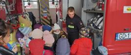 Na zdjęciu widzimy strażaka, który prezentuje grupie dzieci uczestniczącej w zajęciach sprzęt wykorzystywany przez strażaków do gaszenia pożarów.