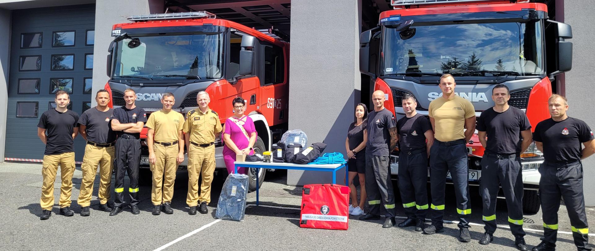 Strażacy i dwie panie w czarnej i fioletowej sukience stoją na tle dwóch samochodów pożarniczych ustawionych w bramach wyjazdowych garażu. Nad nimi napis Państwowa Straż Pożarna. Pomiędzy nimi stolik z drbnymi sprzętami ratowniczymi.
