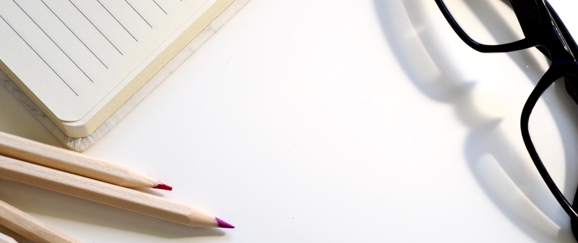 Zdjęcie przedstawiające położone na szkolnym biurku kredki, notes i czarne okulary.
