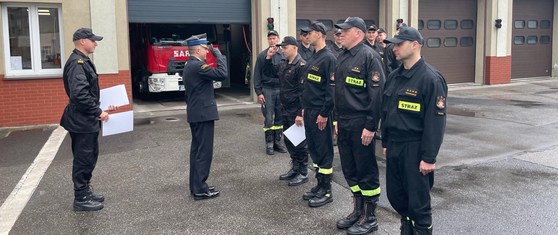 Zdjęcie przedstawia Komendanta Powiatowego PSP w Będzinie wręczającego awanse na wyższe stanowiska służbowe podczas zmiany służby na placu wewnętrznym komendy.