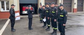 Zdjęcie przedstawia Komendanta Powiatowego PSP w Będzinie wręczającego awanse na wyższe stanowiska służbowe podczas zmiany służby na placu wewnętrznym komendy.
