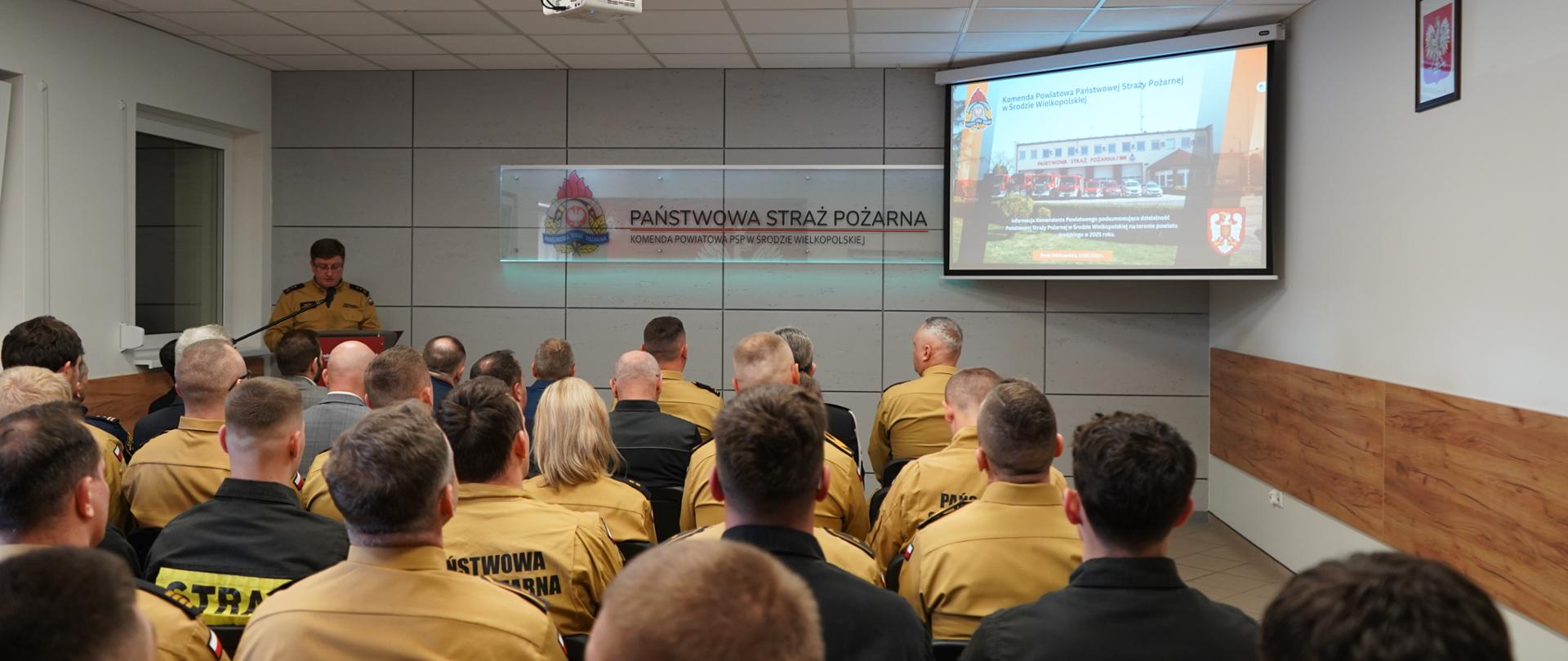 Strażacy siedzą na sali szkoleniowej i oglądają prezentację na odprawie rocznej strażaków