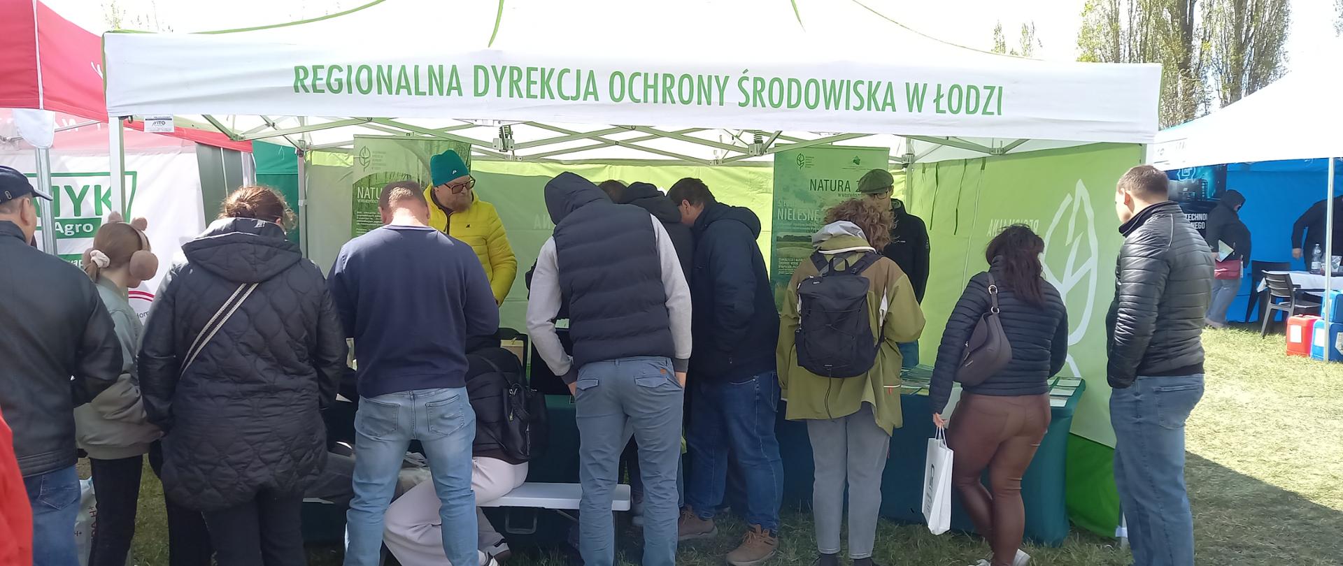 Namiot wystawowy z napisem Regionalna Dyrekcja Ochrony Środowiska w Łodzi. Przed namiotek stoi grupa ludzi ogladjących stoisko. Po bokach widac fragmety innych namiotów. 