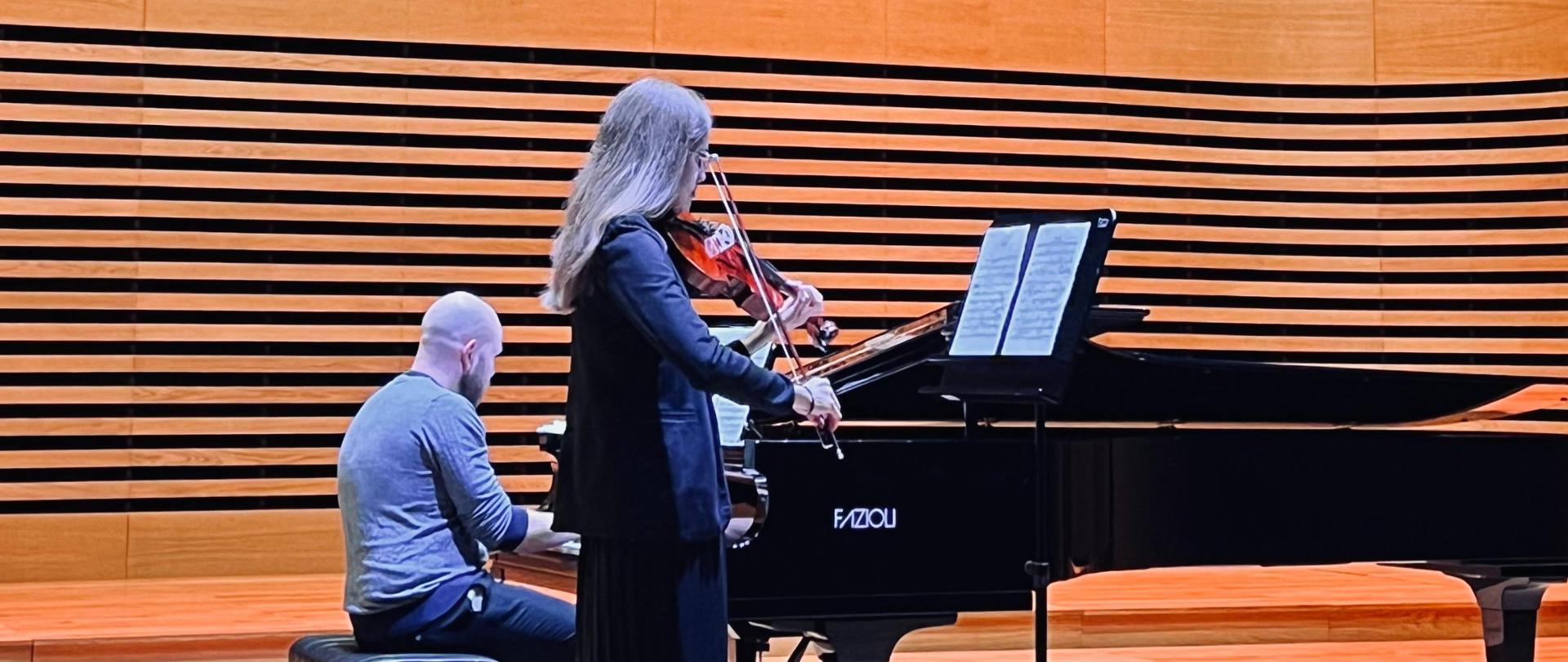 Nastolatka gra na altówce, za nią na fortepianie gra mężczyzna.