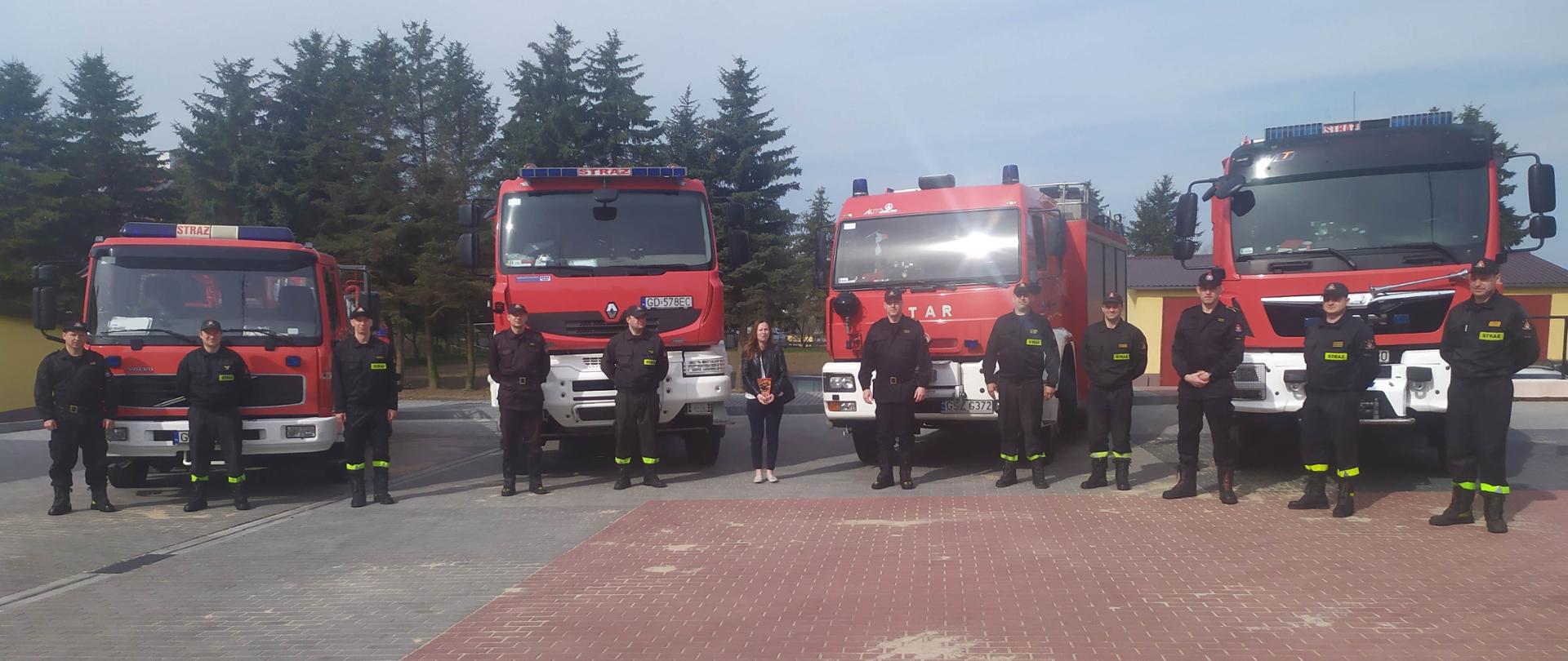 na placu ustawione pojazdy pożarnicze, a przed nimi stoją w odstępach strażacy z JRG Sztum. W środku Pani red. Magdalena Wąsowicz oraz komendant Powiatowy PSP