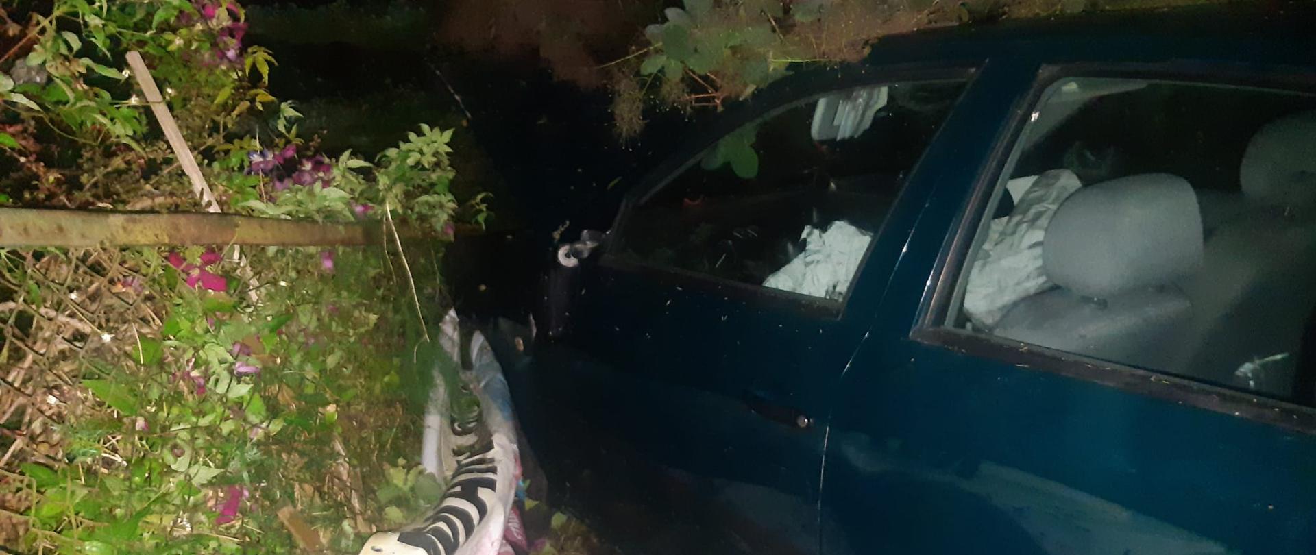 Samochód osobowy stoi w krzakach. Ma uszkodzony przód. Pora nocna. 