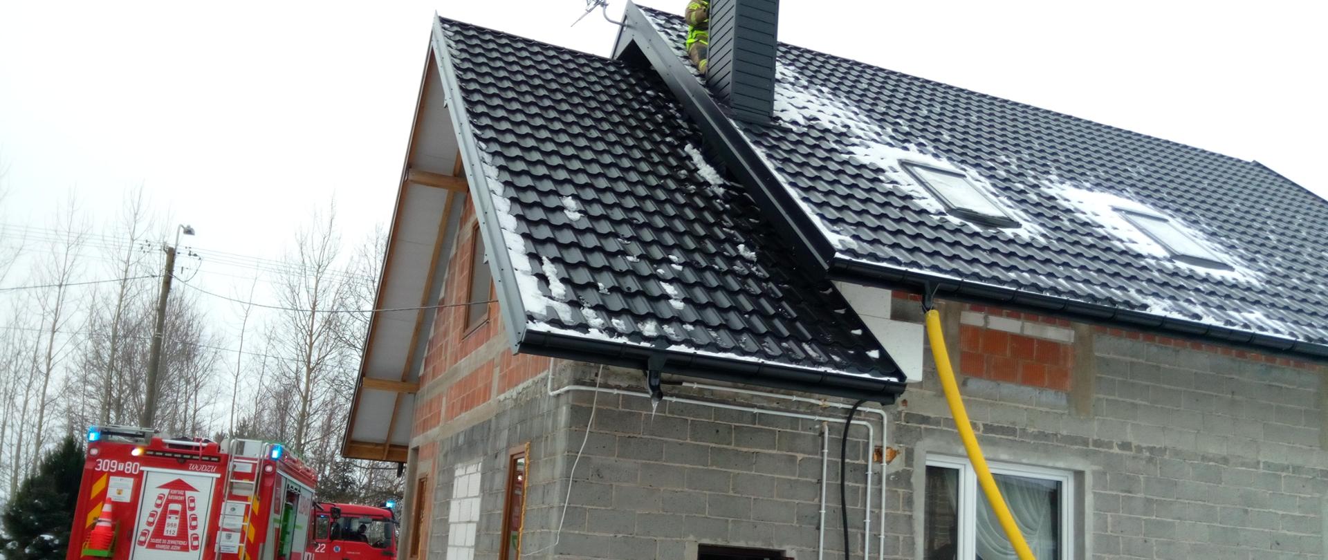 Budynek jednorodzinny w miejscowości Grzymałków przy ulicy Leśniówka 32 od strony podwórza. Na zdjęciu widoczny jest samochód strażacki oraz strażak stojący na dachu za kominem. Nie widać ognia ani dymu.
