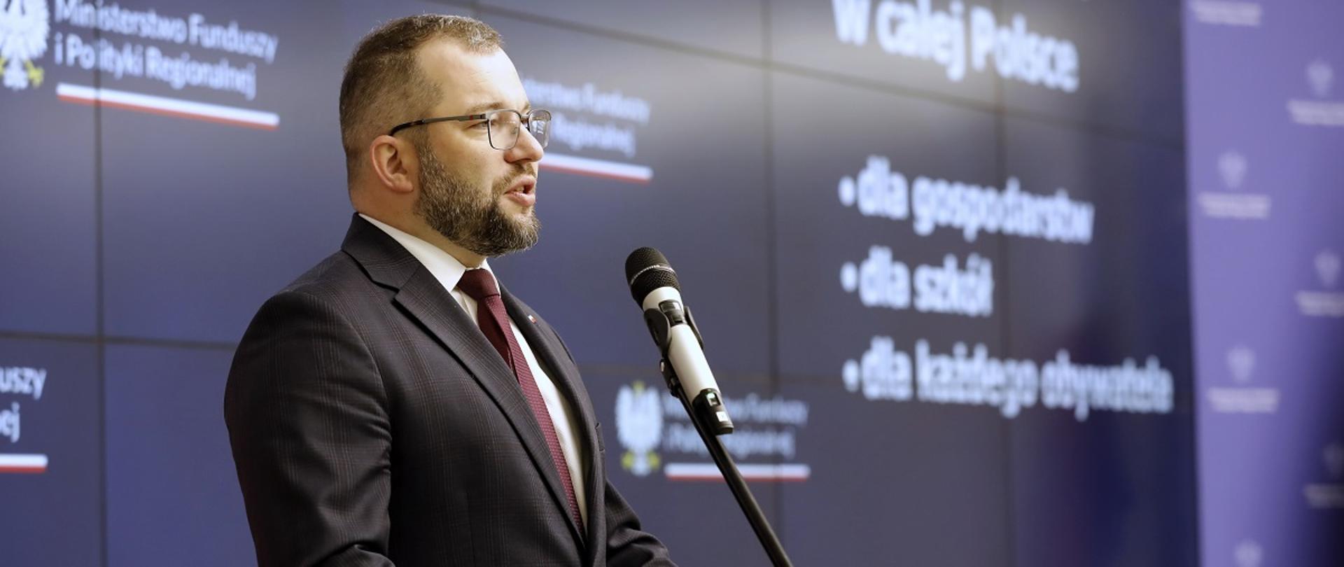 
Minister funduszy i polityki regionalnej Grzegorz Puda przemawia do mikrofonu, za jego plecami baner MFiPR