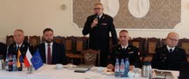 Zdjęcie zrobione w pomieszczeniu. Na pierwszym planie pięć osób za stołem, cztery siedzące oraz jedna stojąca - zastępca śląskiego komendanta wojewódzkiego PSP st. bryg. Rafał Świerczek, który przemawia do mikrofonu.
