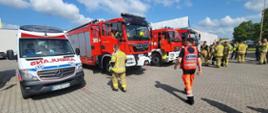 Strażacy stoją obok pojazdów strażackich i karetki pogotowia.
