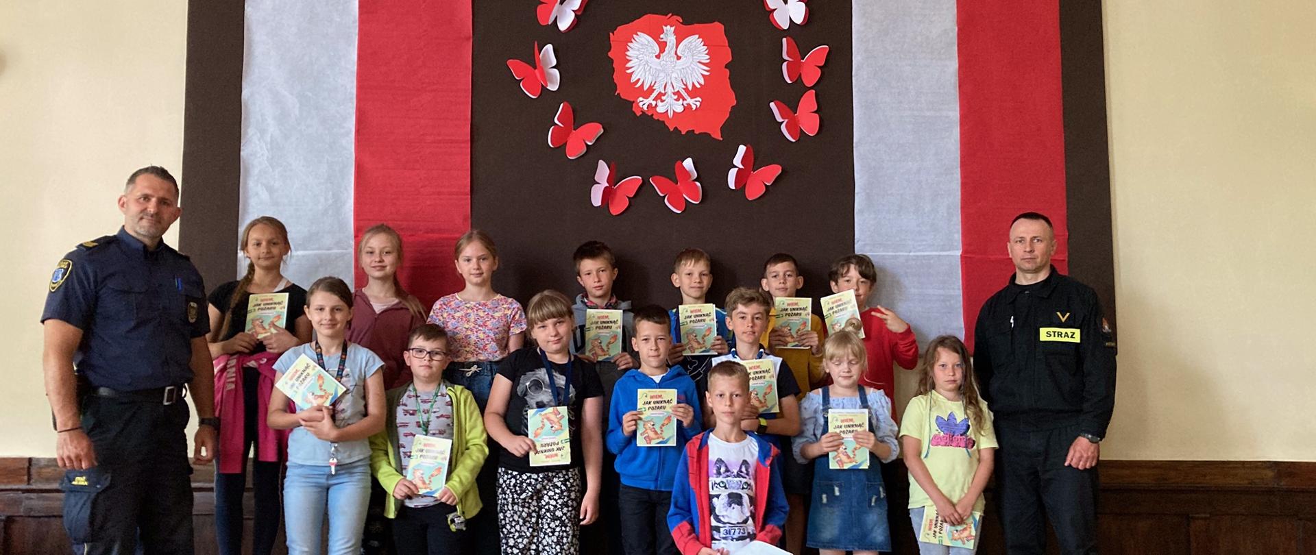 Zdjęcie przedstawia mundurowych: strażaka i strażnika miejskiego w otoczeniu dzieci na tle ścianki z grafiką obrys obszaru polski koloru czerwonego i w środku biały orzeł. Dzieci trzymają w ręku broszurki edukacyjne.
